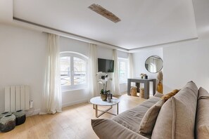 A modern living room with a sofa, a round coffee table, and a TV. There are windows with curtains, a radiator, and a mirror on the wall.