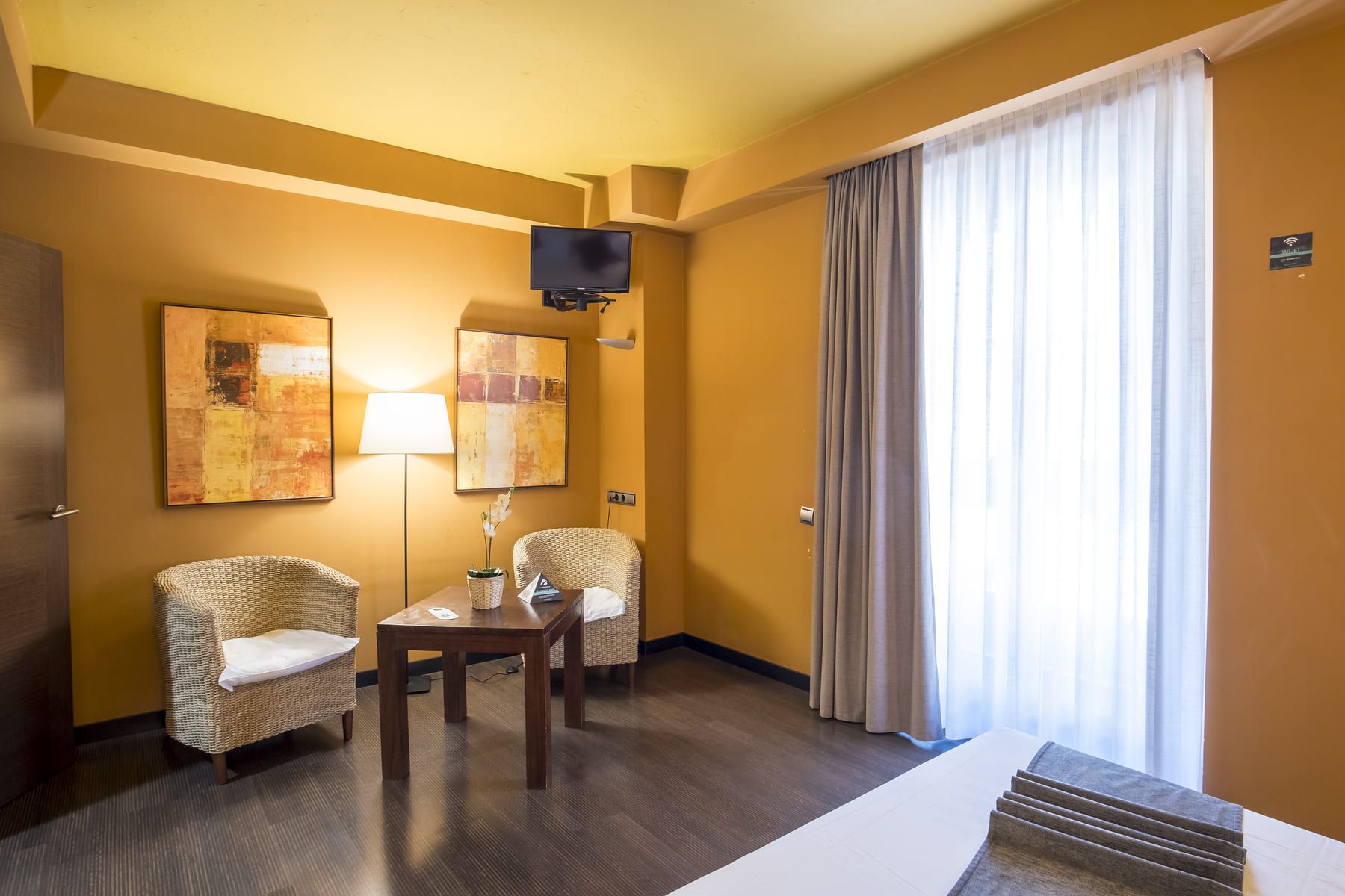 Une chambre d’hôtel avec deux fauteuils en osier, une petite table, un téléviseur à écran plat fixé au mur et une grande fenêtre avec des rideaux.