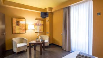 Une chambre d’hôtel avec deux fauteuils en osier, une petite table, un téléviseur à écran plat fixé au mur et une grande fenêtre avec des rideaux.