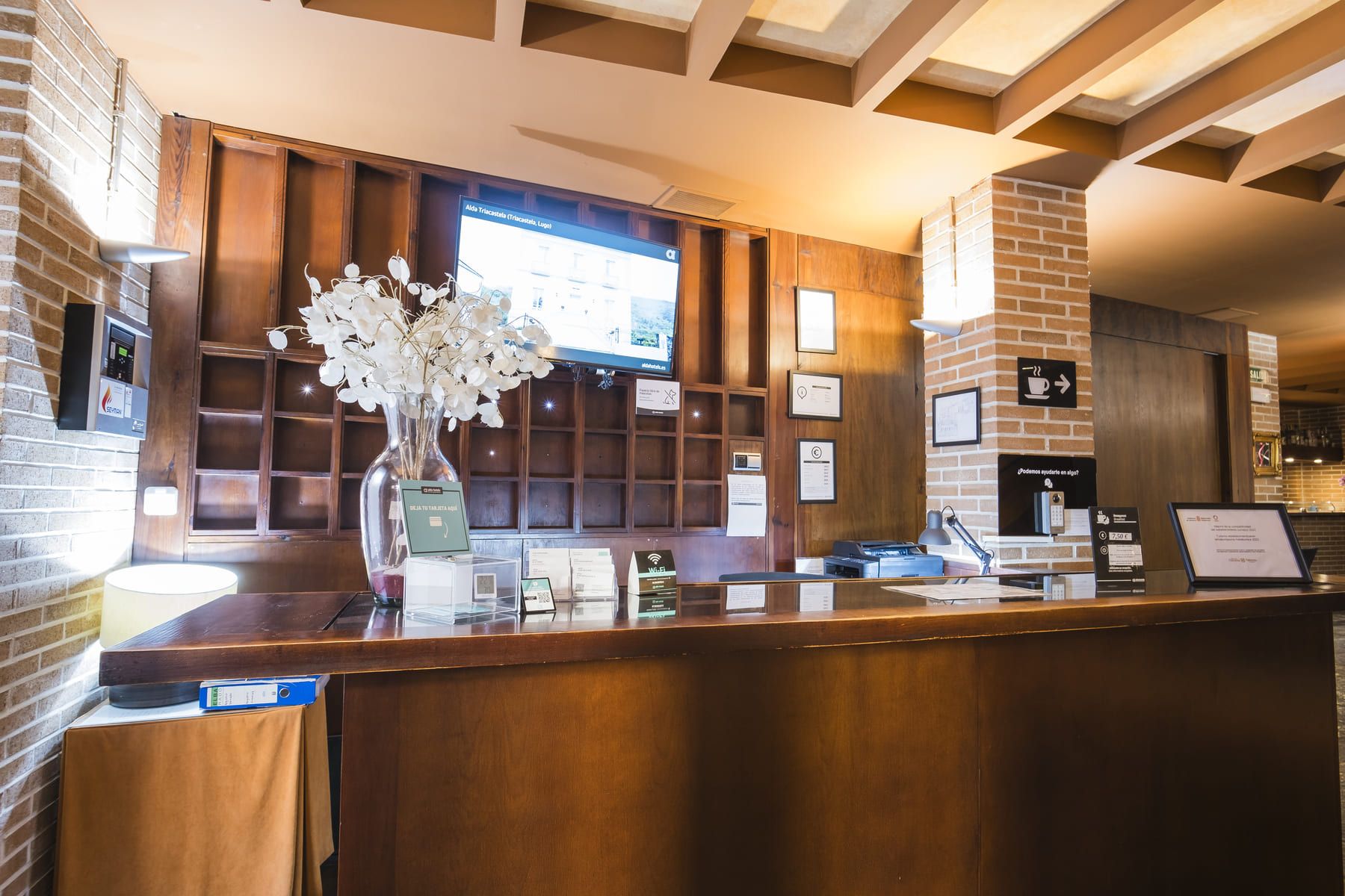 Un mostrador de recepción con encimera de madera, un jarrón con flores blancas, una pantalla digital y un cartel de Wi-Fi.