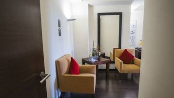A hotel room with a wooden door, two beige armchairs with red cushions, a small wooden table with decorative items, and a large mirror on the wall.