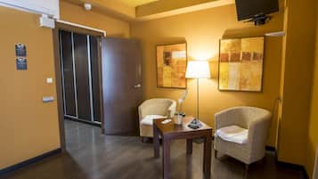 A hotel room with a wooden floor, two wicker chairs, a table with a lamp, and abstract paintings on the wall.