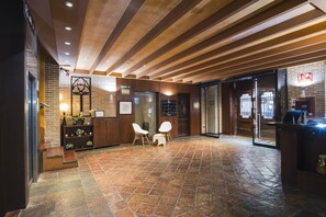 Un hall con techo de madera, paredes de ladrillo, mostrador de recepción, dos sillas blancas y suelo de baldosas.