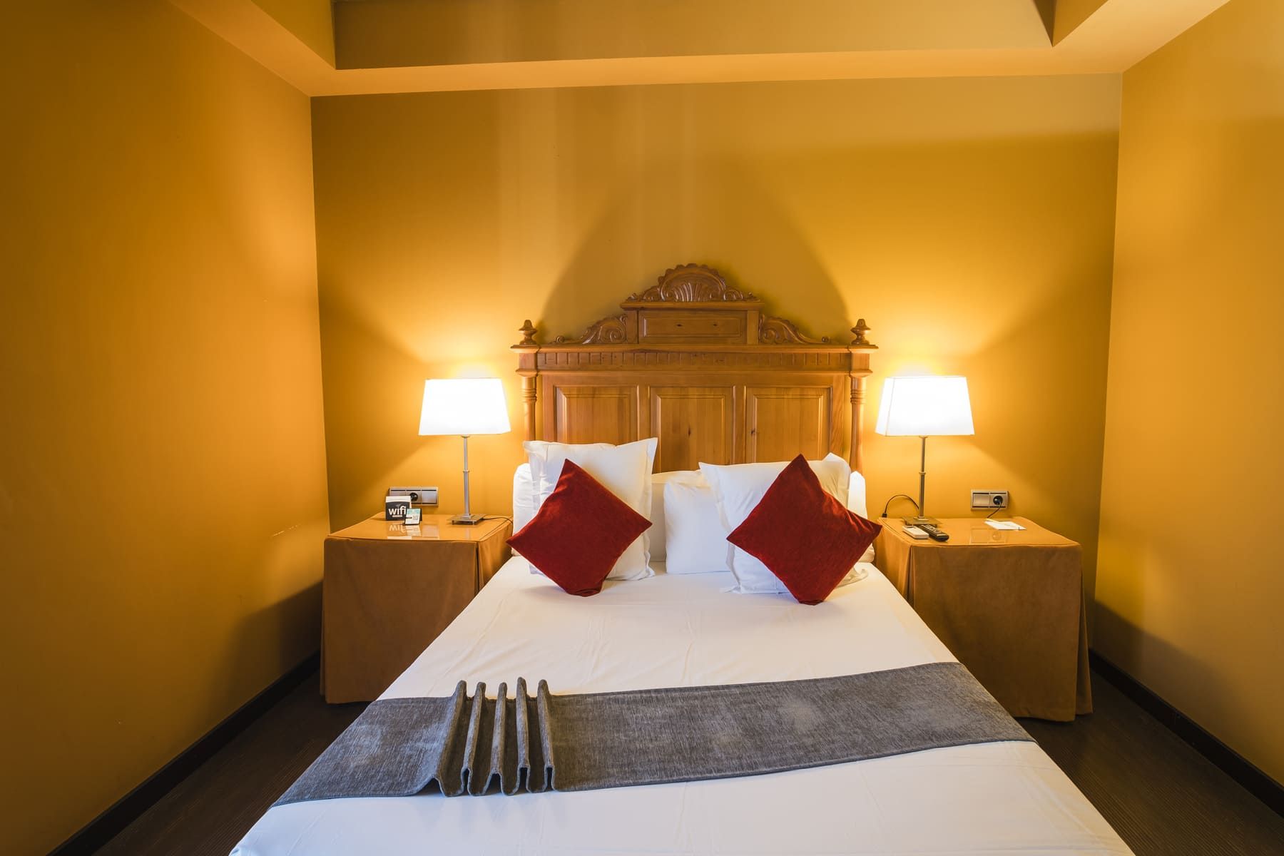Habitación de hotel con una cama grande, dos mesitas de noche, dos lámparas y un cabecero de madera.