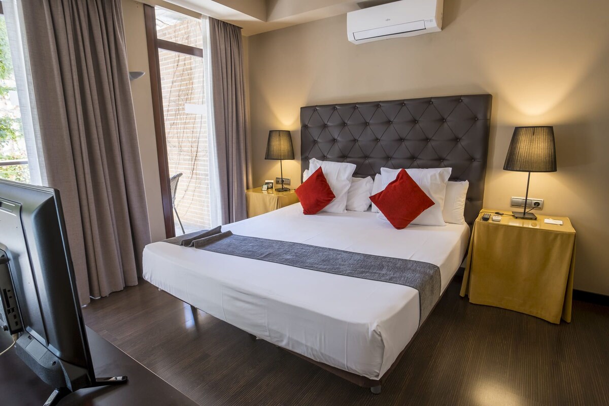 A hotel room with a large bed, two bedside tables with lamps, an air conditioning unit on the wall, and a window with curtains.