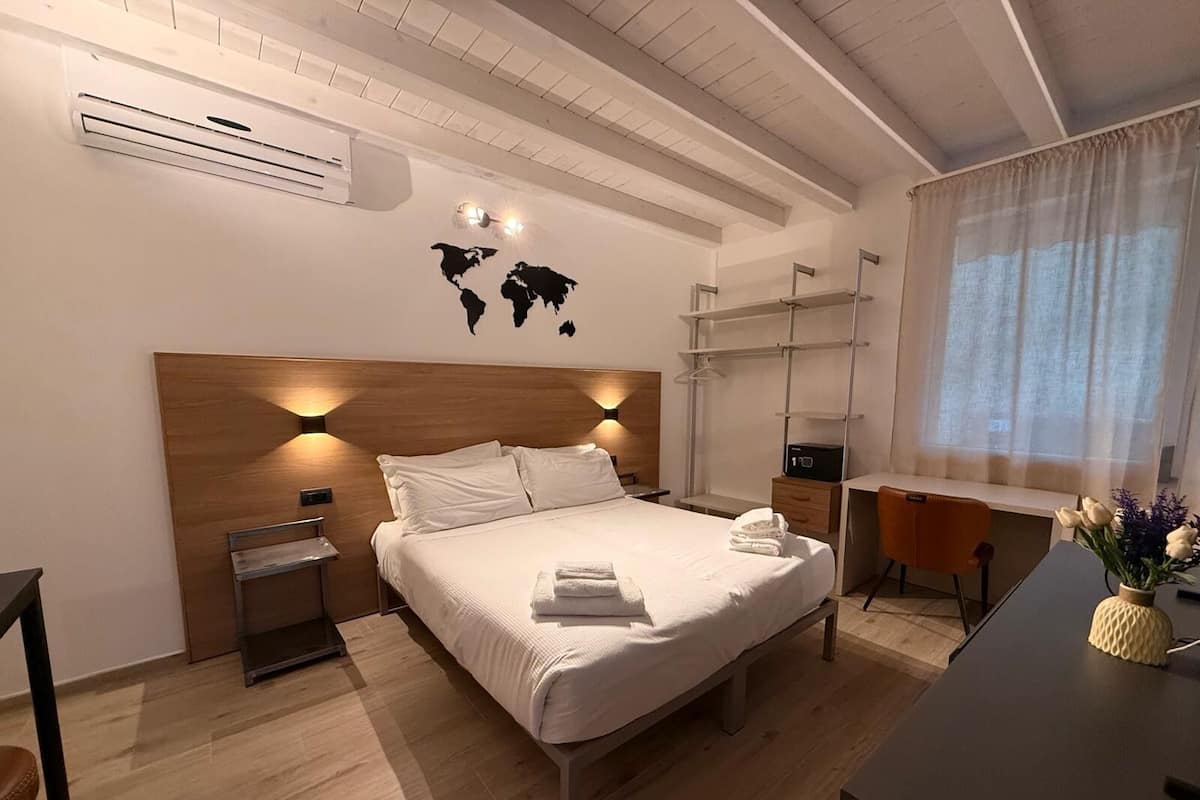 Une chambre d’hôtel moderne avec une tête de lit en bois, un lit avec du linge de lit blanc, un bureau avec une chaise, une petite table de chevet et une carte du monde accrochée au mur.