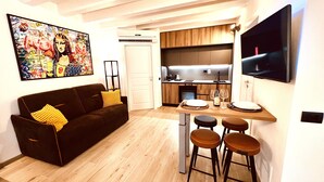 Een compacte leefruimte met een bank, een eettafel met krukken, een keukenhoek en een aan de muur bevestigde televisie.