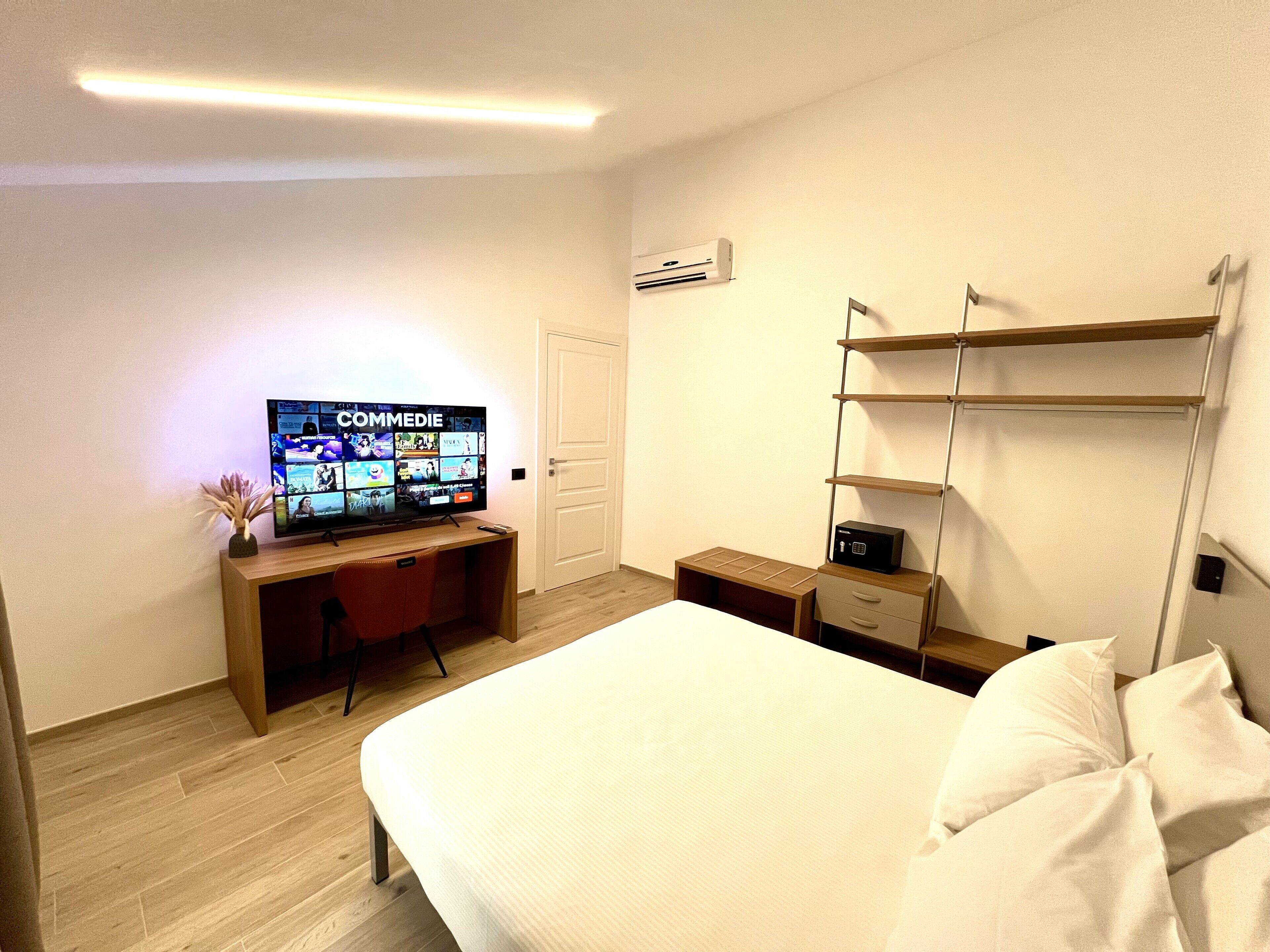 Hotelkamer met een bed, een bureau met een computer, een tv waarop een streamingdienst draait, een plank met een apparaat en een deur.