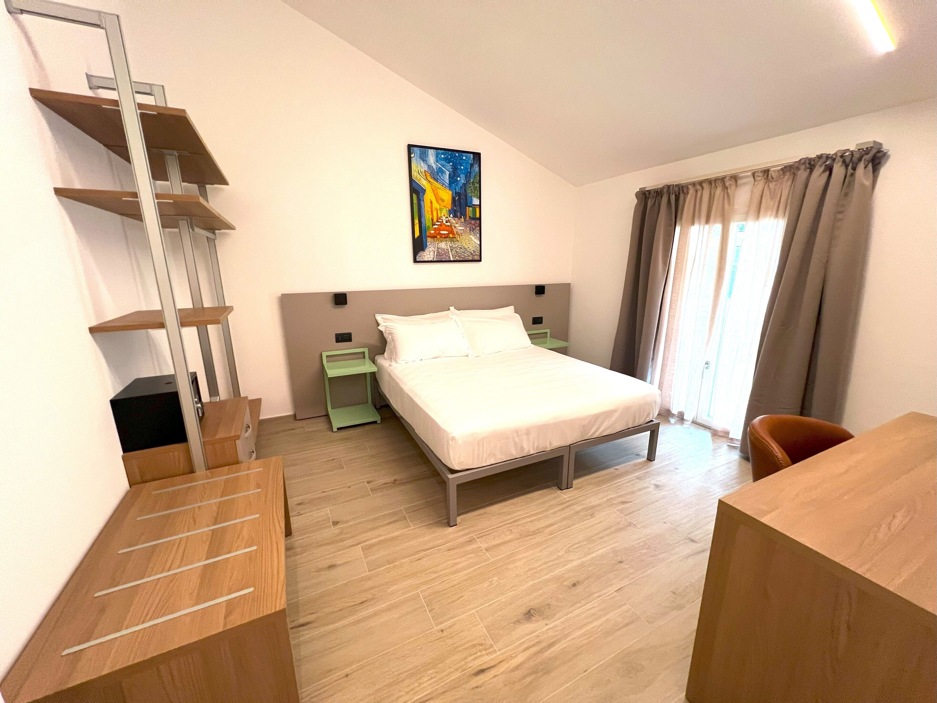 A bedroom with a bed, a nightstand, a green side table, a wooden desk, a chair, a painting on the wall, and a window with curtains.