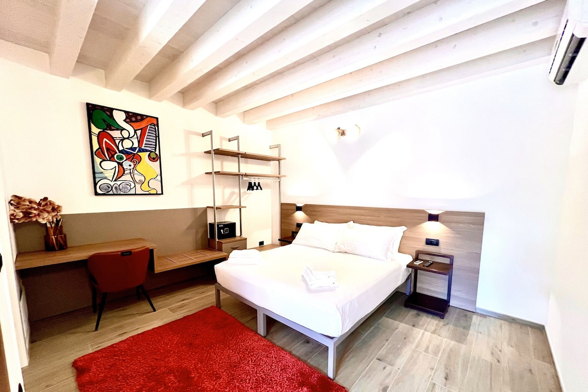 Une chambre d’hôtel moderne, équipée d’un lit, d’un bureau, d’une chaise, d’un tapis rouge et d’une œuvre d’art murale.