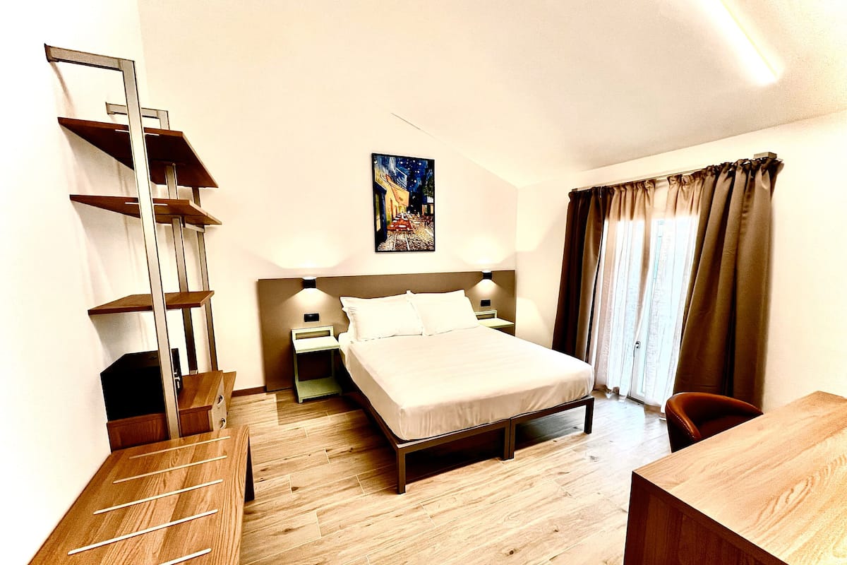 Une chambre d’hôtel avec un lit, un bureau, une chaise, une table de chevet, une lampe, un tableau au mur et une fenêtre avec des rideaux.