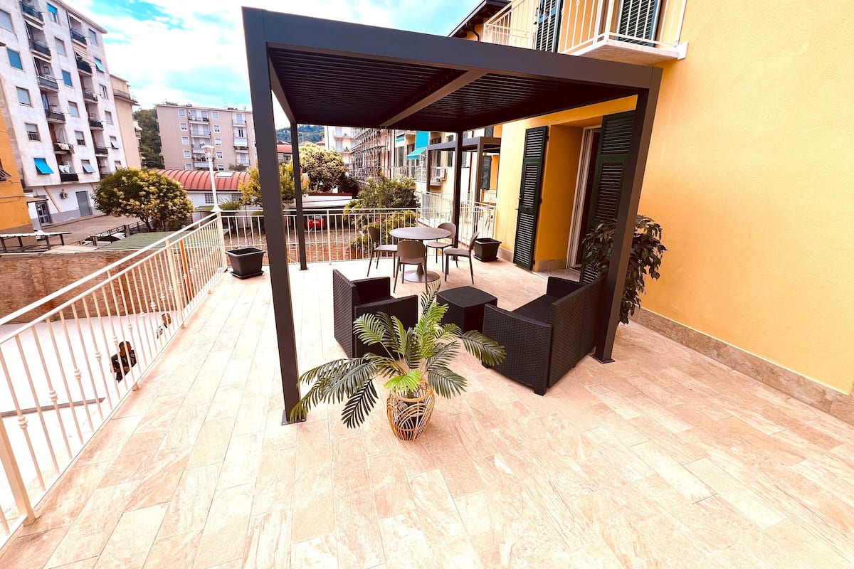 Un espace extérieur aménagé, abrité et doté d’une pergola, de plantes en pot et offrant une vue sur la rue et les bâtiments.