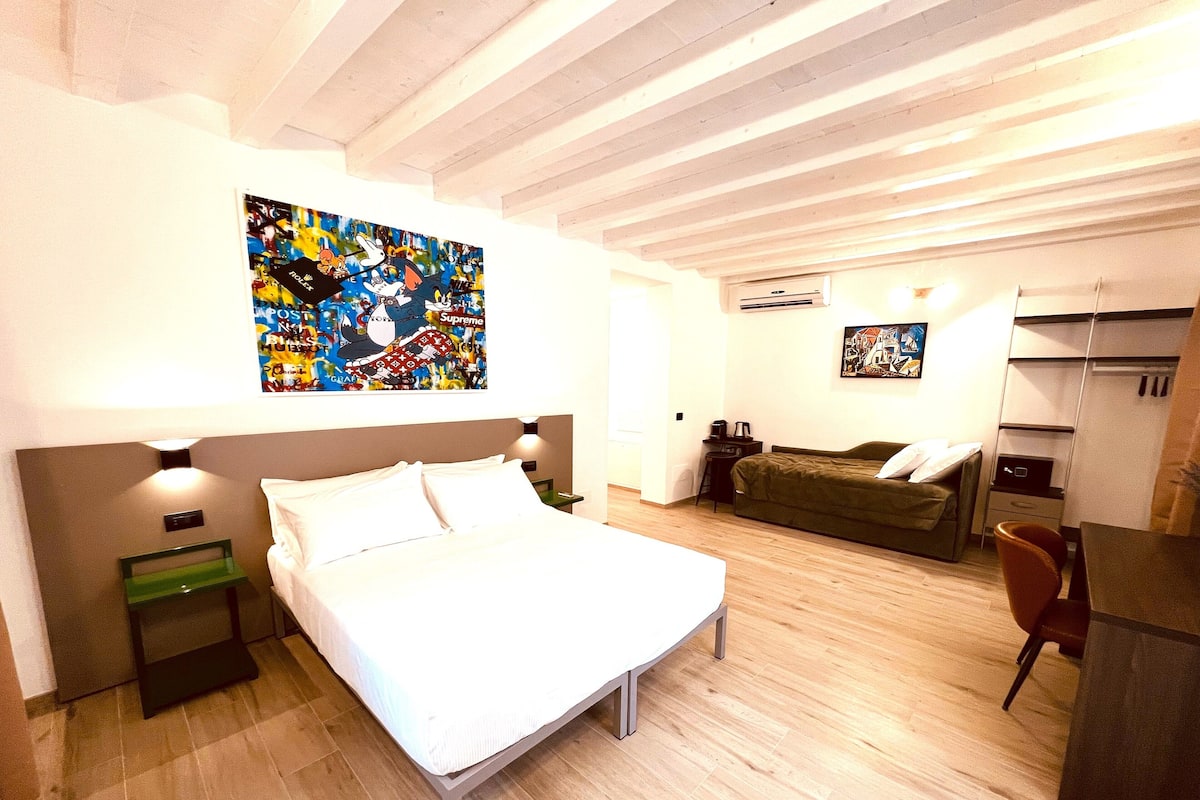Une chambre à coucher avec un lit, un bureau, une chaise et un canapé. Il y a deux tableaux accrochés au mur.