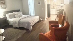 A bedroom with a bed, a chair, and a kitchenette in the background.
