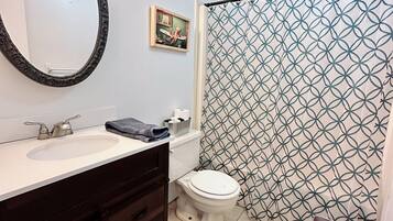 Une salle de bain avec un lavabo blanc, un meuble-lavabo en bois foncé, des toilettes blanches, un rideau de douche à motifs géométriques et une serviette rouge dans un récipient en métal.