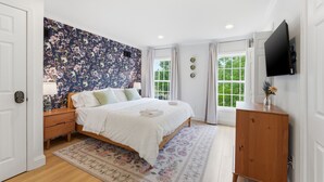 Ein Schlafzimmer mit floralem Wandpapier, einem Bett, einem Nachttisch aus Holz, einer Kommode, einem Fernseher und einem Fenster mit Vorhängen.