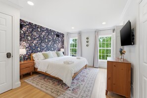 Ein Schlafzimmer mit floralem Wandpapier, einem Bett, einem Nachttisch aus Holz, einer Kommode, einem Fernseher und einem Fenster mit Vorhängen.