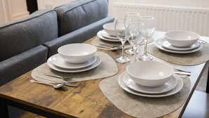 A dining table set with white plates, bowls, and wine glasses, with a grey sofa in the background.