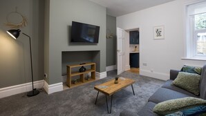 A living room with a flat-screen TV, a wooden coffee table, a grey sofa, and a bookshelf.