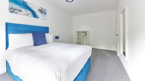 Un dormitorio con una cama grande, cabecera azul, ropa de cama blanca, una pared de acento azul, una lámpara en el techo y una puerta que da a un pasillo.