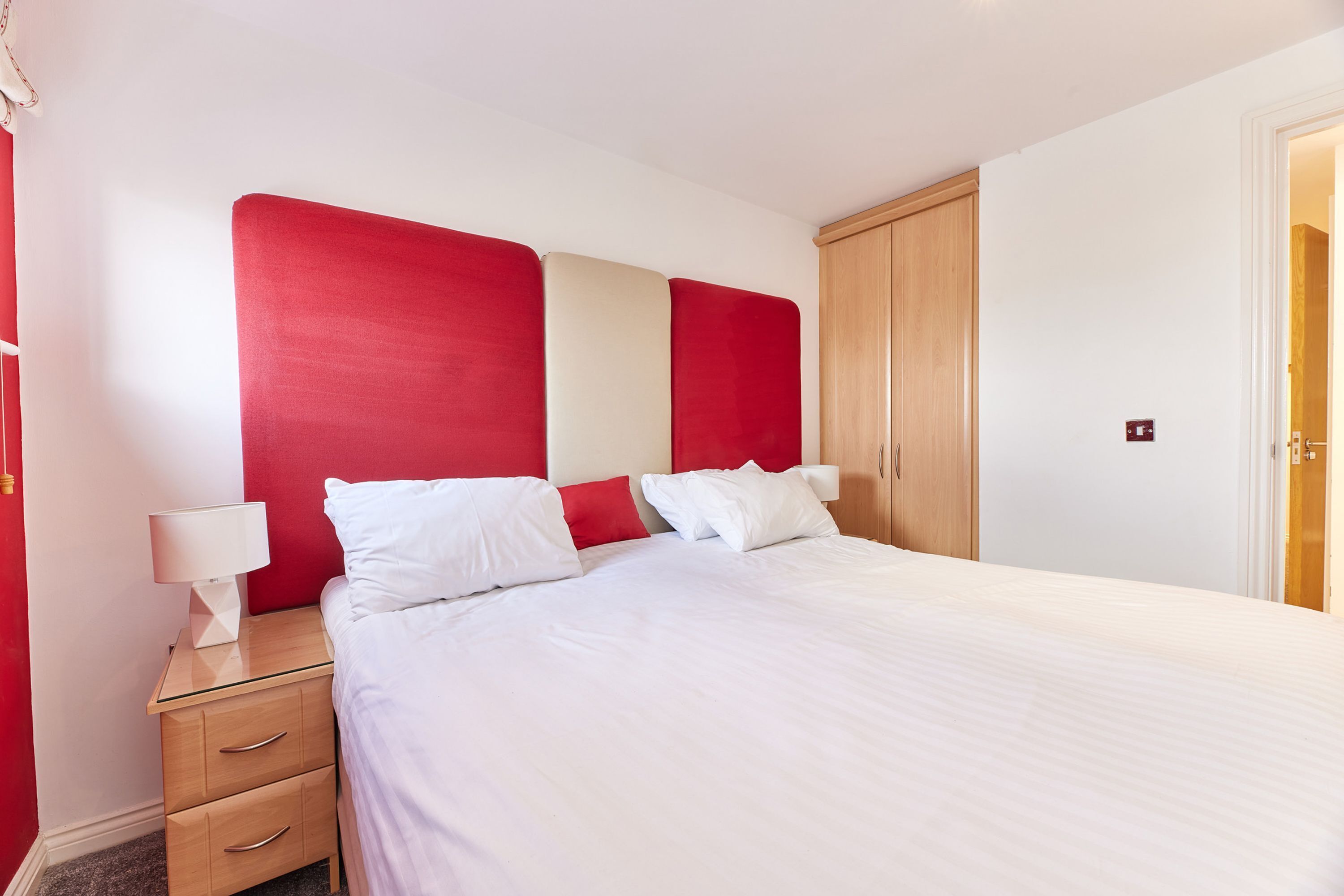 A neatly made bed with white bedding and red headboard accents, a bedside table with a lamp, and a wooden wardrobe.