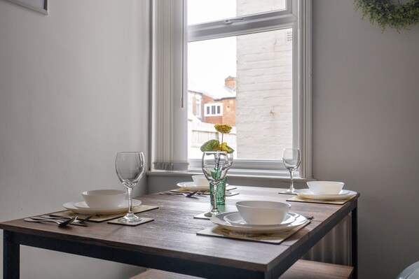 Mesa de comedor dispuesta con cuencos, platos y cubiertos blancos, ubicada cerca de una ventana con vistas a edificios.