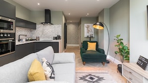 Eine moderne Küche mit Einbauofen und Mikrowelle, ein Essbereich mit einem grünen Sofa und einem gelben Kissen sowie ein Wohnzimmer mit einem grünen Sessel und einer Stehlampe.