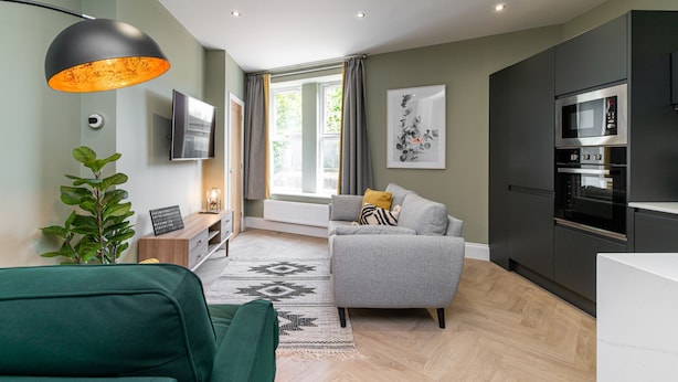 Ein modernes Wohnzimmer mit grauem Sofa, grünem Sessel, einer schwarzen Pendelleuchte, einem Flachbildfernseher und einer Küchenzeile mit Mikrowelle und Backofen.