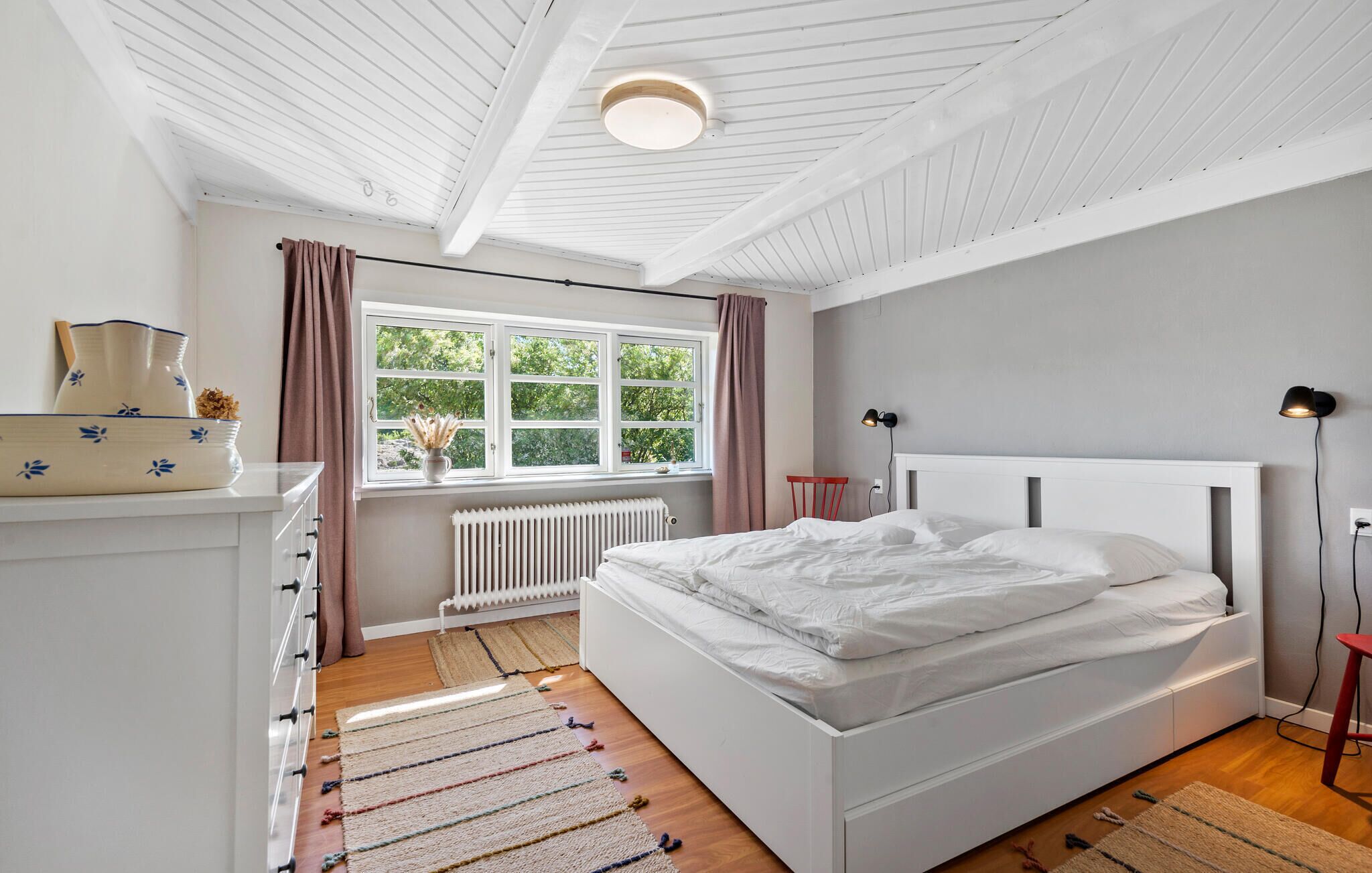 A bedroom with a white bed, a radiator, a window with curtains, a dresser, and a lamp.