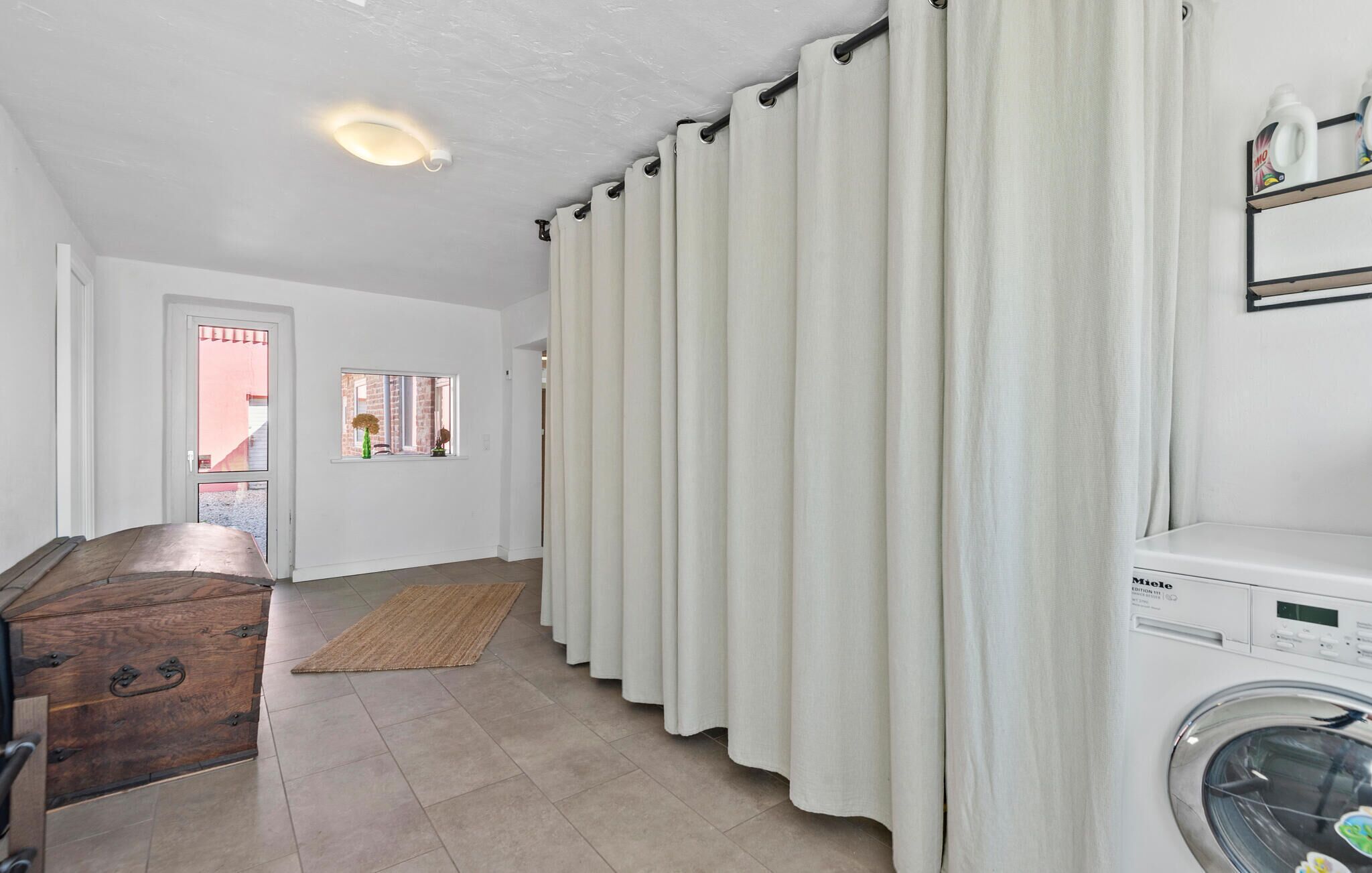 A laundry room with a washing machine, a wooden chest of drawers, a rug, and a window with a view of a building.
