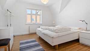 A bedroom with a sloped ceiling, a bed with white bedding, a desk with a chair, a standing lamp, and a window with a view of a tiled roof. - Nice home in Løgstør with WiFi (Løgstør)