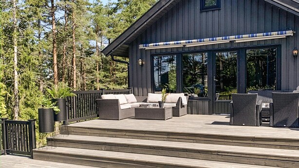 Une maison moderne dotée d’une terrasse, de mobilier d’extérieur et d’une grande fenêtre donnant sur une forêt.