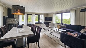 Un salon spacieux avec une grande table à manger, des sièges confortables, une cheminée et de grandes fenêtres donnant sur l’extérieur.