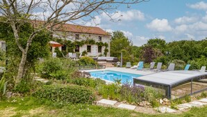 Une piscine entourée de chaises longues, un bâtiment en pierre avec un toit en tuiles et une végétation luxuriante.