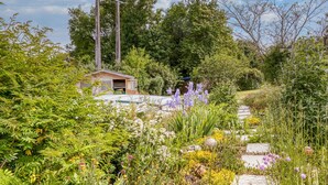Un jardin avec un petit abri en bois, un chemin de pierres et diverses plantes et fleurs.