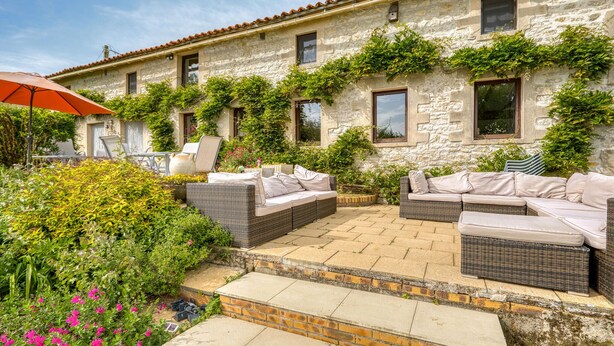 Un bâtiment en pierre doté d’un patio aménagé avec des meubles en osier et un parasol rouge.