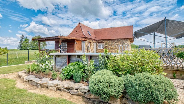 A rustic stone house with a red roof, a wooden porch, and a garden with various plants and a stone wall. - Superbe maison piscine privée (Payzac)