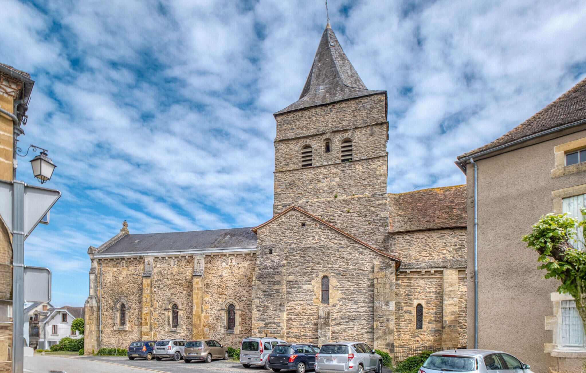 A historic stone building with a prominent tower and arched windows, surrounded by parked cars and adjacent buildings.
