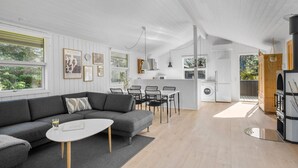 A modern living room with a gray sofa, a small white table, and a dining area with chairs. There is a kitchen area with a refrigerator and a washing machine.