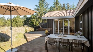 An outdoor patio with a table and chairs, a large umbrella, and a wooden deck leading to a covered area with a tiled roof.