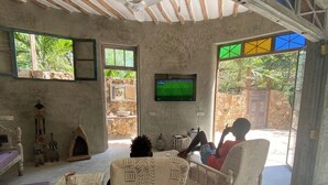 A rustic living room with a television, two patterned chairs, a small table, and a stone fireplace. - Tulua House on Galu Beach: Private Villa @ Tamani (Diani)