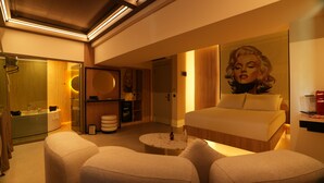 Quarto de hotel moderno com uma cama grande, área de estar com duas cadeiras, mesa de centro redonda, banheiro com box de vidro e armário de madeira.