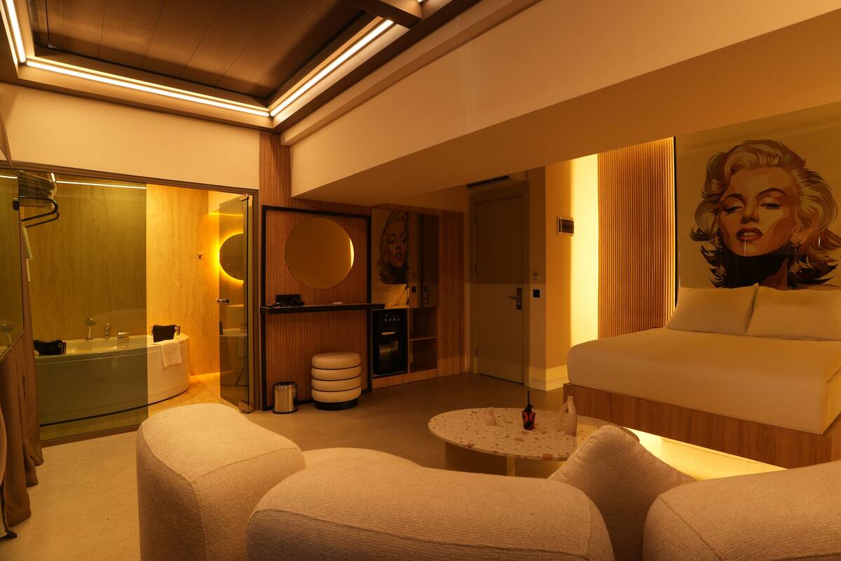 A modern hotel room with a large bed, a sitting area with two chairs, a round coffee table, a bathroom with a glass shower enclosure, and a wooden wardrobe.