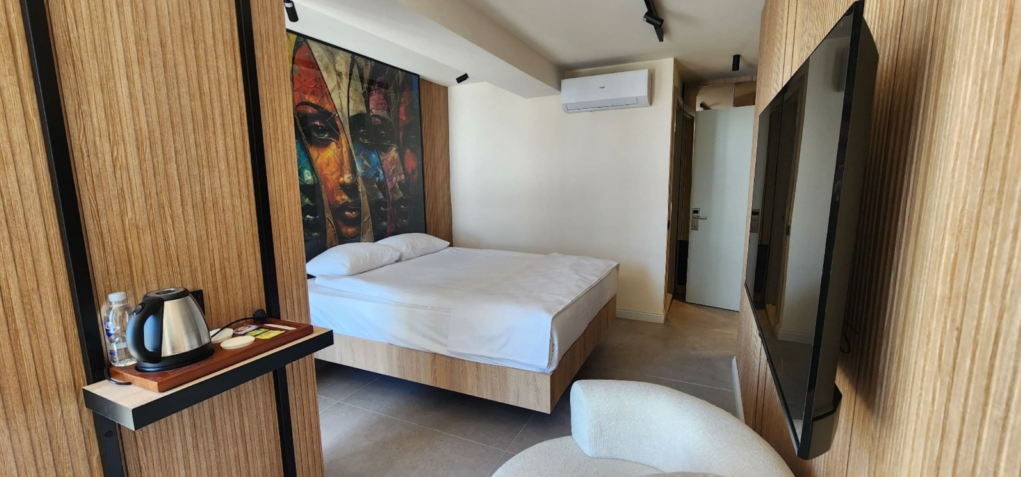 A modern hotel room with a bed, a painting on the wall, a TV, and a small table with a kettle and water bottle.