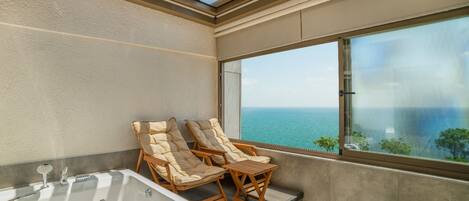 A balcony with a large window overlooking the sea, two cushioned chairs, and a bathtub.