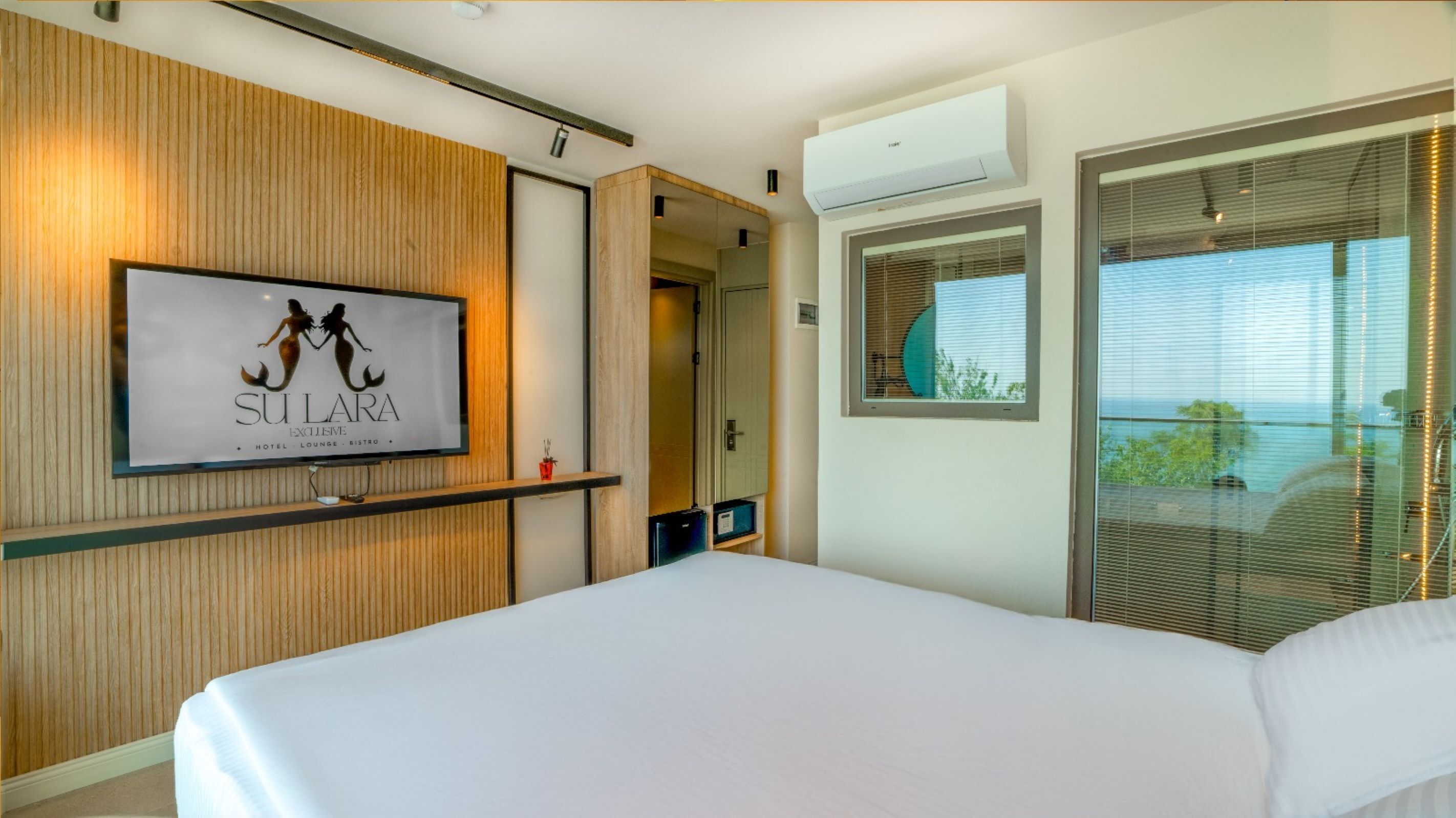 A hotel room with a large bed, a mounted TV displaying 'SULLARA EXCLUSIVE HOTEL LOUNGE BISTRO', wooden paneling, an air conditioning unit, and a balcony with a view.