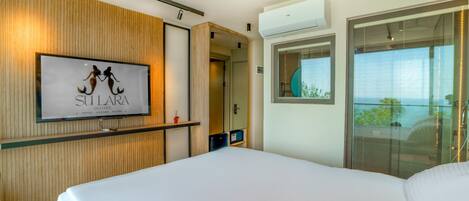 A hotel room with a large bed, a mounted TV displaying 'SULLARA EXCLUSIVE HOTEL LOUNGE BISTRO', wooden paneling, an air conditioning unit, and a balcony with a view.
