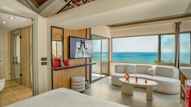 Quarto de hotel moderno com uma grande janela oferecendo vista para o mar, um sofá, uma mesinha e uma TV fixada na parede.
