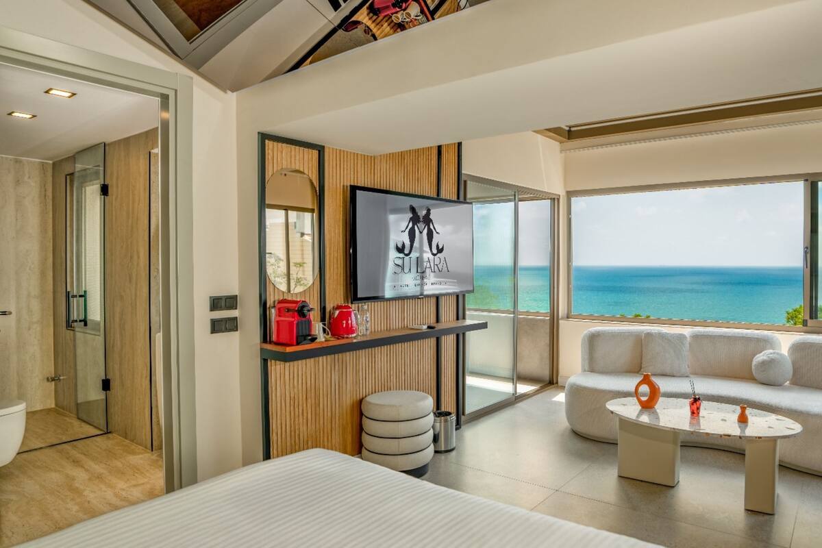 A modern hotel room with a large window offering a view of the sea, a sofa, a small table, and a TV mounted on the wall.