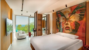 Um quarto moderno com uma cama grande, um espaço de estar com uma poltrona e uma mesa, uma janela ampla com vista para o oceano, e um quadro abstrato colorido na parede.